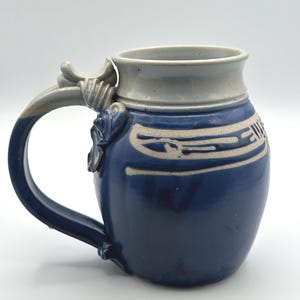 May include: A blue ceramic beer stein with a light gray rim and handle. The stein features a decorative design in a lighter color around the body. The handle has a decorative flourish near the top. The mug is designed for holding beverages.
