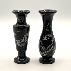 Könnte beinhalten: Zwei schwarze Steinvasen mit geätzten weißen Blumen- und Landschaftsdesigns. Die Vasen haben eine ausgestellte Ober- und Unterseite mit einem schlanken, länglichen Körper. Eine Vase ist höher und kantiger, die andere runder.