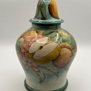 May include: A ceramic jar with a lid, featuring a hand-painted design of fruits like pears, grapes, and peaches. The jar has a green base, lid, and accents, with a light blue background. The jar is approximately 10 inches tall.
