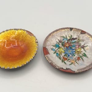 May include: Two antique enamel dishes. One is oval-shaped with a yellow and orange interior and a beaded edge. The other is round, featuring a floral design in blue, yellow, and red on a white background with a silver rim.