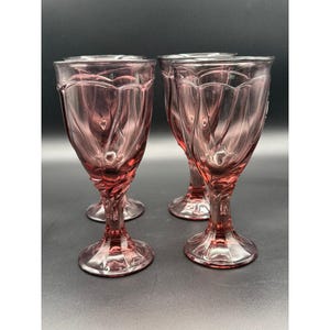 May include: A set of four translucent pink glass goblets featuring a molded tulip-like floral pattern on the bowl and a twisted stem design. Each glass has a circular base and a smooth rim, showcasing a consistent rose-tinted hue throughout.