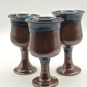 Puede incluir: Tres cálices de cerámica con acabado brillante, que presentan un cuerpo de color marrón rojizo intenso y un esmalte azul oscuro en contraste alrededor del borde acampanado y la base. Cada recipiente tiene un cuenco redondeado y un pie circular.
