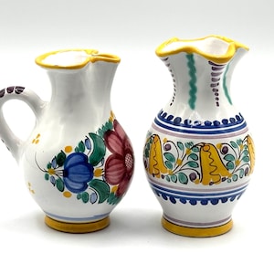 May include: Two white ceramic pitchers with floral designs. One pitcher has a handle and a red, blue, and green floral pattern. The other has a yellow, blue, and green floral pattern. Both have yellow rims and bases.