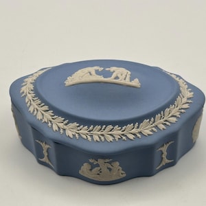 May include: A blue ceramic trinket box with a decorative lid featuring a white relief of two figures. The box has a white leaf garland around the lid and sides, with additional relief designs. A classic decorative item.