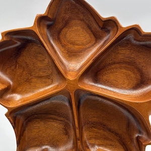 Puede incluir: Una bandeja de madera para servir con forma de hoja, con cinco compartimentos. La bandeja está hecha de madera marrón oscura y está soportada por un soporte de metal negro. Los compartimentos están diseñados para contener aperitivos.