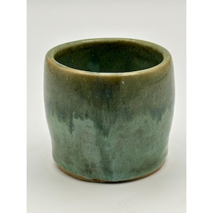 Op de afbeelding: Een handgemaakte keramische beker met een afgeronde vorm. De buitenkant heeft een mix van groene en bruine tinten, met een glanzende afwerking. De bovenrand heeft een donkerdere, bruinachtige tint, terwijl de body overgaat in een lichter groen. De beker is geschikt voor dranken.
