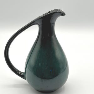 Könnte beinhalten: Eine dunkelgrüne Keramikkrug mit einem schwarzen Griff und Ausguss. Der Krug hat eine glatte, glänzende Oberfläche und eine Tropfenform. Der Griff verläuft elegant von oben nach unten.