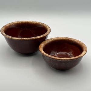 May include: Two brown ceramic bowls with a glossy finish and contrasting tan rims. The bowls are of different sizes, with a larger bowl behind a smaller one. Ideal for serving food or as decorative items.