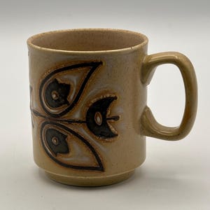 May include: A tan ceramic mug featuring a black and brown floral pattern. The mug has a curved handle and a speckled finish. The design is symmetrical and stylized, giving it a vintage aesthetic. The mug is suitable for hot drinks.