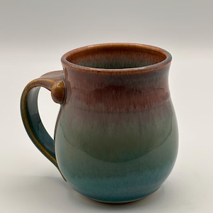 Pode incluir: Uma caneca de cerâmica artesanal com um corpo arredondado e uma alça grande. A caneca apresenta um gradiente de cores, passando de uma borda marrom escura para uma base turquesa. A alça tem um detalhe dourado.