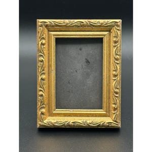 May include: A rectangular picture frame with a metallic gold finish and an ornate, raised scrollwork pattern along the border. The frame features a dark, solid-coloured interior backing and a stepped, multi-layered profile design.