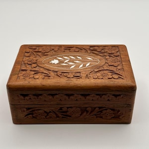 May include: A rectangular wooden box with intricate floral carvings. The box is a warm brown color, with a lighter oval-shaped inlay on the lid featuring a leaf design. The box is likely used for storing jewelry or small keepsakes.
