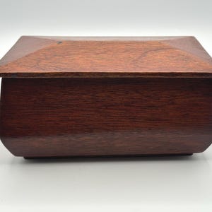 May include: A rectangular wooden box with a dark reddish-brown finish. The box has a slightly angled lid and rounded edges, giving it a classic and elegant appearance. The wood grain is visible, adding to its natural aesthetic.