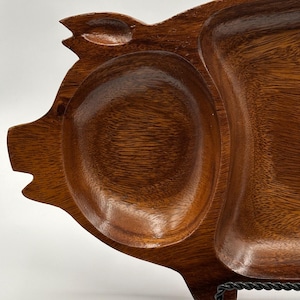Puede incluir: Una bandeja de madera con forma de cerdo. La bandeja tiene tres compartimentos: un óvalo grande, un óvalo más pequeño y una sección rectangular. La madera tiene un color marrón cálido y un patrón de vetas visible. Soporte de metal negro.