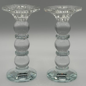 Könnte beinhalten: Zwei klare Glaskerzenhalter. Jeder hat einen runden Sockel, einen Stiel mit drei kugelförmigen Abschnitten und eine ausgestellte Oberseite. Die Oberteile haben ein Sternmuster. Das Glas hat einen leichten blau-grünen Farbton.