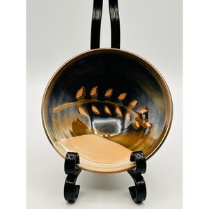 Pode incluir: Uma tigela de cerâmica artesanal com um desenho de folhas. A tigela apresenta um esquema de cores azul escuro e castanho com um interior bege. O desenho das folhas é em tons de castanho e bege. A tigela é exibida em um suporte de metal preto.