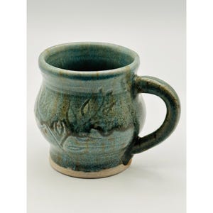 Peut inclure: Mug en céramique fait à la main avec un glaçage turquoise et un motif de feuilles. La tasse a un corps arrondi, une anse robuste et une base de couleur claire. La tasse mesure environ 10 cm de haut et 9 cm de diamètre.