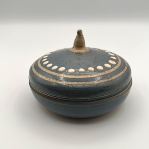 May include: A round, lidded ceramic jar with a speckled blue-grey glaze. The lid features a central pointed handle, a ring of white dots, and concentric beige circular lines. The base is a matching blue-grey bowl with a smooth, rounded profile.