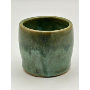 May include: A cylindrical ceramic cup featuring a reactive glaze with blended shades of sage green, forest green, and earthy brown. The vessel has a smooth, rounded rim and a slightly tapered base, showcasing a glossy finish with subtle color variations.