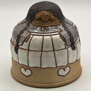 Könnte beinhalten: Keramiktopf mit Deckel, geformt wie eine Person mit dunklen Zöpfen. Der Topf ist weiß mit einem Gittermuster, und der Sockel ist beige mit zwei Herzformen. Der Deckel hat ein lächelndes Gesicht.