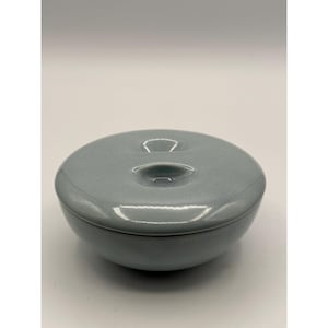 May include: A round, light blue ceramic bowl with a matching lid. The lid features two small, recessed, oval-shaped indentations on its top surface. The bowl has a smooth, glossy finish and a simple, circular silhouette with a uniform color throughout.