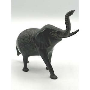May include: A dark bronze-colored metal elephant figurine standing with its trunk raised. The elephant features intricate, embossed floral and geometric patterns across its back and sides, with a textured surface and four sturdy legs.