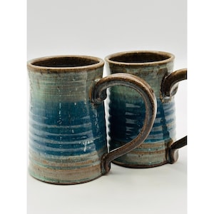 Pode incluir: Duas canecas de cerâmica feitas à mão com esmalte azul e verde. As canecas têm formato cilíndrico com alça e borda marrom escuro. As canecas têm aproximadamente 13 cm de altura.