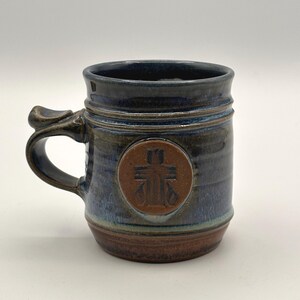 May include: A handcrafted ceramic mug with a dark blue glaze, a brown base, and a handle. The mug features a circular emblem with a cross design. The mug is approximately 4 inches tall.
