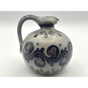 May include: A round ceramic pitcher with a light gray base and a dark blue glazed pattern. The design features swirling scrollwork, circular motifs, and small teardrop shapes. It has a single curved handle and a small spout at the top rim.