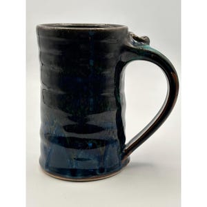 Peut inclure: Une grande tasse en céramique cylindrique avec un vernis brillant sombre aux tons bleu marine profond et noir. La surface présente une texture subtilement ondulée sur le corps et comprend une anse incurvée fixée sur le côté avec un accent turquoise.