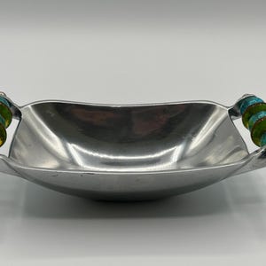 Könnte beinhalten: Eine Servierschale aus gebürstetem Aluminium mit dekorativen Griffen. Die Griffe weisen einen Stapel aus grünen, türkisfarbenen und gelblichen Perlen auf. Die Schale hat eine rechteckige Form mit sanft geschwungenen Seiten und einem leicht konkaven Boden.