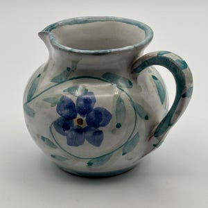 May include: A small, round ceramic pitcher with a handle and spout. The pitcher is white with blue floral and green leaf designs. The rim and handle are accented with a teal-colored trim. The pitcher is likely used for serving liquids.