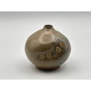 May include: A round ceramic vase with a narrow neck and a smooth, glossy finish in shades of tan, brown, and gray. The surface features a diagonal row of six small, raised circular floral impressions and subtle horizontal brushstroke textures.