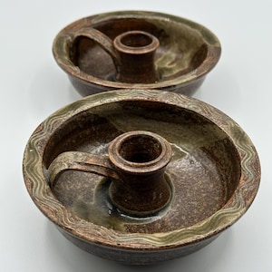 Op de afbeelding: Twee handgemaakte keramische kandelaars. Elk heeft een cirkelvormige basis met een handvat en een centrale beker voor de kaars. Het aardewerk is bruin met groene en beige accenten. De rand heeft een golvend ontwerp.