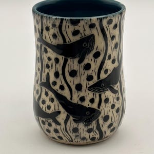 Pode incluir: Um copo de cerâmica com exterior creme com ilustrações de baleias pretas e interior azul escuro. O design inclui pontos pretos e linhas onduladas, criando uma cena subaquática. O copo é cilíndrico com uma parte superior ligeiramente alargada.