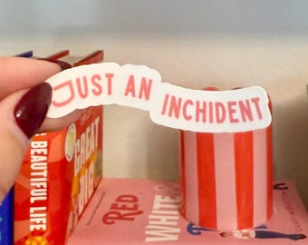 May include: A shelf displays a collection of books with colorful spines. The books include titles like "Iron Flame" and "Fourth Wing." A sticker with the text "Just an Incident" is held in front of a striped container.