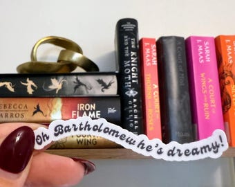 Op de afbeelding: Een hand die een witte sticker vasthoudt met de tekst "Oh Bartholomew he's dreamy!" in een speels lettertype. De sticker bevindt zich voor een boekenplank vol kleurrijke boeken, waaronder titels als "Iron Flame" en "A Court of Thorns and Roses".