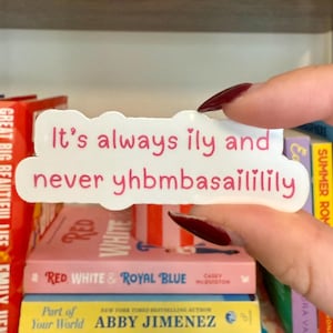 May include: A white sticker with pink text that reads "It's always ily and never yhbmbasaililily." The sticker is held in front of a bookshelf filled with colourful books.