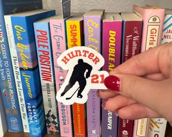 Puede incluir: Una pegatina con la silueta negra de un jugador de hockey, el nombre "Hunter" en rojo y blanco y el número "21". La pegatina se sostiene frente a una estantería llena de libros coloridos.