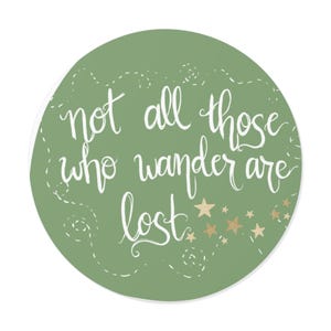 May include: A circular, sage green sticker featuring the quote "not all those who wander are lost" in white cursive script. Gold stars and dotted lines embellish the design. The sticker is set against a white backdrop.