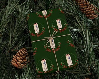 May include: A wrapped gift with dark green wrapping paper featuring a repeating pattern of a man in a military uniform, the words "Mr. Darcy", and the phrase "All I want for Christmas is". The gift is tied with twine and set on a bed of pine needles and pine cones.