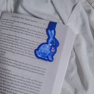 May include: A blue bookmark shaped like a rabbit, featuring a constellation design and a crescent moon. The bookmark is placed in a book with the title "EMPIRE OF THE DAWN" visible.