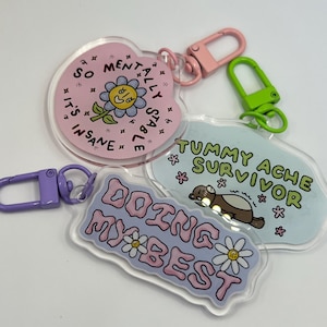 May include: A collection of acrylic keychains with various designs and phrases. One keychain is pink and reads "So Mentally Stable, It's Insane" with a flower graphic. Another says "Tummy Ache Survivor" with a cartoon animal. The last says "Doing My Best".