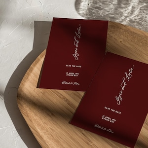 May include: Two rectangular deep red save the date cards featuring white text that reads Joyce and Leslie, Save the Date, 17 April 1971, Byron Bay, and Details to Follow. The cards are positioned on a light-colored wooden surface with shadows.
