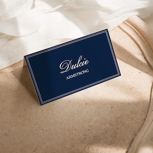 Peut inclure: Un marque-place rectangulaire bleu marine plié avec un double cadre blanc. Le prénom Dulcie est imprimé en police cursive blanche au-dessus du nom ARMSTRONG en lettres capitales blanches sans empattement au centre du panneau avant.