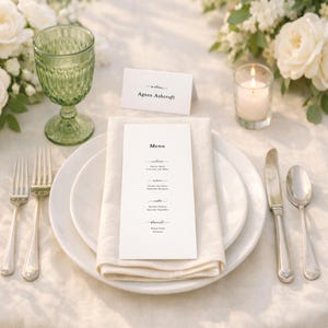 Könnte beinhalten: Elegante Tischdekoration mit weißen Tellern, Besteck und einer gefalteten Serviette. Eine Menükarte und eine Namenskarte sind auf dem Teller platziert. Ein grünes Glas und eine brennende Kerze sind ebenfalls vorhanden, zusammen mit weißen Blumen. Die Menükarte lautet "Menu".