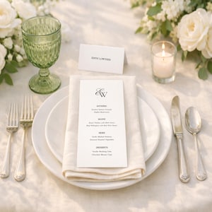 Peut inclure: Mise en place élégante avec assiettes blanches, couverts et un menu. Un verre vert et une bougie allumée sont présents. Le menu affiche les initiales "E&W" et une liste de plats. Des roses blanches et de la verdure ajoutent une touche naturelle.