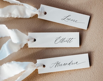 Könnte beinhalten: Drei rechteckige weiße Papieranhänger mit runden Löchern, jeweils mit einem ausgefransten weißen Seidenband versehen. Die Anhänger tragen die Namen Lenore, Elliott und Alexandria in einer eleganten, schwarzen Schreibschrift.