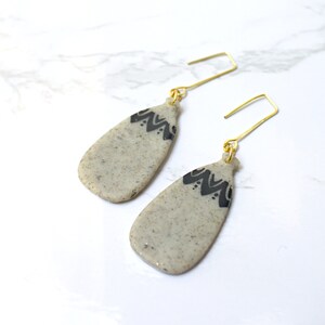 May include: A pair of dangle earrings with a teardrop shape. The earrings are a speckled beige color with a black geometric design at the top. The ear wires are gold-colored.