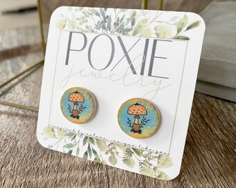 May include: A pair of round wooden stud earrings featuring a mushroom design. The earrings have a blue and yellow background with a brown mushroom and red spots. The card reads "POXIE jewelry".